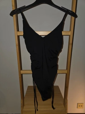 Svart baddräkt med dragsko - Snygg svart baddräkt med djup V-ringning och smala axelband. Dragsko-detaljer på sidorna ger en trendig och justerbar passform. Perfekt för stranden eller poolen. Materialet känns stretchigt och följsamt.