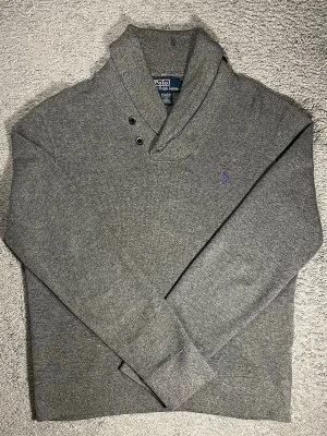 Grå stickad tröja Polo Ralph Lauren - Snygg grå stickad tröja från Polo Ralph Lauren med lila broderad logga på bröstet. Tröjan har en unik sjalkrage med två metallknappar och långa ärmar. Perfekt för dig som gillar klassisk och stilren design. 