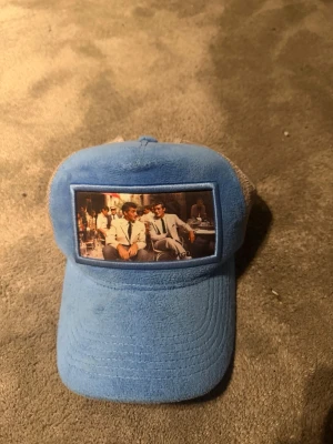 Blå meshkeps med tryck framtill - Snygg blå keps med mesh på sidorna och bak, samt ett tryck framtill med motiv av personer i kostym. Kepsen har en böjd skärm och justerbar passform. Märket Mads är broderat på sidan. Perfekt för dig som vill sticka ut med en unik accessoar.