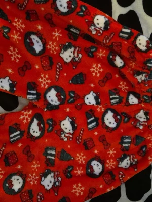 Hello Kitty julpyjamasbyxor röd - Mysiga röda pyjamasbyxor med juligt Hello Kitty-motiv, snöflingor, julgranar, presenter och polkagrisar. Byxorna har resår i midjan och är gjorda i mjukt fleece-material, perfekta för vinterkvällar.