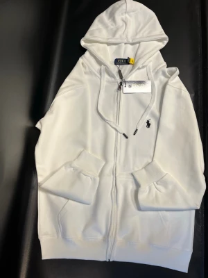 Vit hoodie från Polo Ralph Lauren - Snygg vit hoodie från Polo Ralph Lauren med dragkedja framtill, två fickor och klassisk huva med dragsko. Ikoniska lilla svarta loggan broderad på bröstet. Tillverkad i mjukt bomullsmaterial som känns riktigt skönt mot huden. Perfekt för en clean och stilren look.