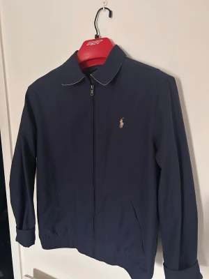 Marinblå vindjacka från Ralph Lauren - Klassisk marinblå vindjacka från Ralph Lauren med dragkedja framtill och broderad logga på bröstet. Jackan har en enkel krage med kontrastfärg på insidan, ribbade muddar och nederkant samt två sidofickor. Perfekt för en stilren och avslappnad look.