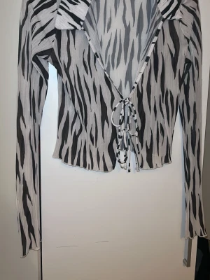 Zebramönstrad topp med knyt - Snygg zebramönstrad topp med långa ärmar och knyt framtill. Toppen har en djup v-ringning och är gjord i ett lätt, ribbat material som känns luftigt. Perfekt för dig som vill sticka ut med ett djuriskt mönster och trendig look.