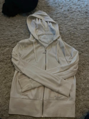 Vit zip hoodie med guldvingar - Säljer en vit hoodie från Victoria's Secret med dragkedja och huva. På ryggen finns snygga guldglittriga änglavingar och framtill ett litet logotryck. Hoodien har fickor och ribbade muddar. Perfekt för dig som gillar en clean men ändå unik stil.