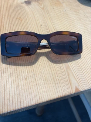 Bruna solglasögon Vogue - Snygga solglasögon från Vogue med rektangulära bågar i mörkbrunt. Glasen är tonade och bågarna har en klassisk, stilren look. Perfekta för dig som vill ha en trendig accessoar med en touch av retro.