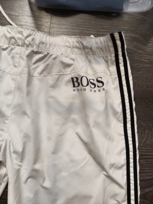 Vita mjukisbyxor från B Helg REA NU! - Säljer ett par vita mjukisbyxor från  B med svarta dubbla ränder längs sidorna och elastisk midja. Byxorna har en avslappnad passform och är perfekta för en chill och sportig look. B-logga tryckt på vänster sida. Materialet känns mjukt och bekvämt. Är stor m 