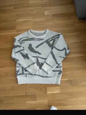  stickad tröja stolek M manfinty  - Säljer en ljusgrå stickad tröja med abstrakt mörkgrått mönster. Tröjan har rund halsringning, ribbade muddar och nederkant. Perfekt för dig som gillar en chill och modern stil. Manfinity