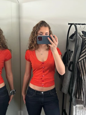 Röd croppad topp med knappar - Snygg röd croppad topp från New Look med korta ärmar och v-ringning. Toppen har ribbade kanter och dekorativa knappar framtill. Perfekt till jeans eller kjol för en trendig look.