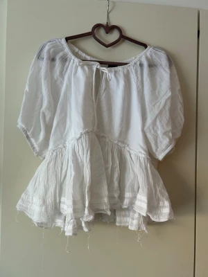 Vit volangblus med knytning - Luftig vit blus med volanger och korta puffärmar. Blusen har en rundad halsringning med knytband och är tillverkad i ett tunt, strukturerat bomullsliknande tyg. Perfekt för en avslappnad och trendig look.