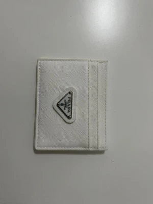 Vit kortfodral från Prada - Snyggt vitt kortfodral från Prada i canvasmaterial med det klassiska triangulära metalloggan framtill. Fodralet har flera kortfack och en stilren, minimalistisk design som passar perfekt i fickan. Perfekt accessoar för dig som gillar lyxiga detaljer.