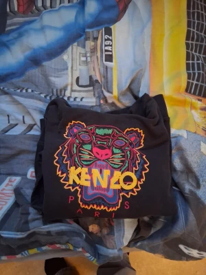 Svart Kenzo hoodie med tigertryck (Lãs beskrivning) - Säljer en svart hoodie från Kenzo med ikoniskt färgglatt tigerbroderi och texten 'KENZO PARIS' på bröstet. Hoodien har huva och stor magficka framtill. Perfekt för dig som vill sticka ut med en streetwear-look. Storleken är stor på mig som är 180 och det är därför jag säljer
