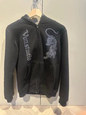 Svart Vonstudio hoodie med tigertryck - Säljer en svart hoodie från Vonstudio med dragkedja, huva och fickor. På framsidan finns ett stort tigertryck i silvriga stenar samt Vonstudio logga i liknande stil. Hoodien har en avslappnad passform och är tillverkad i mjukt material, perfekt för en cool streetwear-look. Och alla stenar sitter kvar 