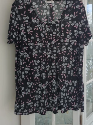 Blommig svart topp från Generous - Svart topp från Generous med blommigt mönster i vitt och rosa. Toppen har korta ärmar, rundad halsringning och knappar framtill. Materialet känns mjukt och luftig. 