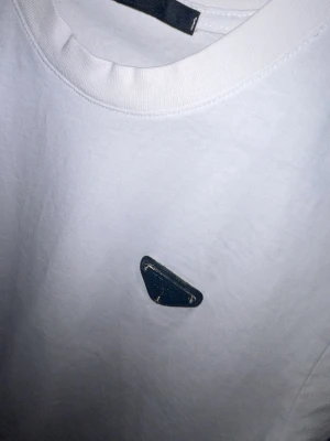 Vit t-shirt med metallmärke Prada  - Vit t-shirt i bomull med rund hals och korta ärmar. På bröstet sitter ett litet metallmärke i triangelform som ger en clean och modern vibe. Perfekt till jeans eller shorts för en enkel men snygg look.