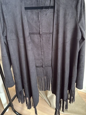 Svart fransad cardigan från Zara Basic - Snygg svart cardiganjacka från Zara Basic i mjukt mockaimitationsmaterial. Öppen modell utan knäppning och långa ärmar. Jackan har coola fransar längs nederkanten som ger en boho-vibe. Perfekt att slänga över en t-shirt eller topp för en avslappnad look.