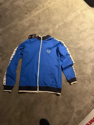 Blå track jacket från Fred Perry - Snygg blå track jacket från Fred Perry med vita detaljer och svart ribbad mudd. Dragkedja framtill, vita ränder med logga längs ärmarna och broderad Fred Perry-logo på bröstet. Perfekt för en sportig och trendig look.