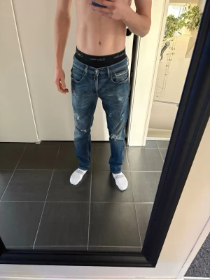slitna replay jeans  - Säljer ett par blå jeans från Replay med snygga slitningar och tvättade detaljer. Jeansen har raka ben, klassiska fem fickor och dragkedjegylf. Perfekta för en avslappnad och trendig look. Materialet är denim i bomull.