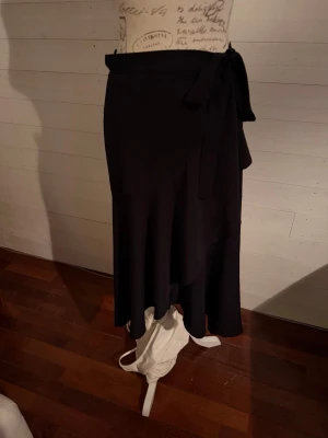 Svart omlottkjol med knytband - Stilren svart omlottkjol med knytband i midjan. Kjolen har en mjuk och följsam passform och faller snyggt längs benen. Perfekt för dig som gillar minimalistisk och tidlös stil.