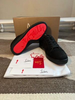 Svarta sneakers från Christian Louboutin - Snygga svarta sneakers från Christian Louboutin med klassisk röd sula och diskret logga. Skorna har en låg profil, svarta skosnören och är tillverkade i mocka och läder. Perfekta för dig som vill ha en exklusiv och stilren look.