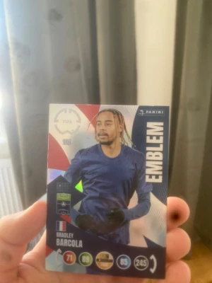 Panini FIFA spelkort Bradley Barcola - Säljer ett Panini FIFA spelkort med Bradley Barcola. Kortet har färgstark design i blått, rött och vitt med texten 'EMBLEM' på sidan. Perfekt för samlare eller dig som gillar fotbollsspel och trading cards.