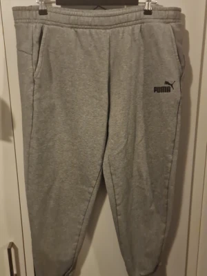 Grå mjukisbyxor från Puma - Säljer ett par klassiska grå mjukisbyxor från Puma med resår i midjan och muddar vid bensluten. Byxorna har två sidofickor och Puma-logga tryckt på vänster ben. Perfekta för chill eller träning. Materialet är mjukt och skönt, troligtvis bomullsblandning.