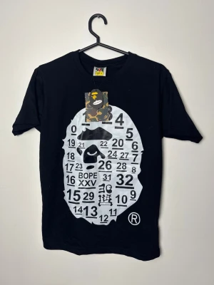 Svart BAPE t-shirt med tryck - Svart t-shirt från BAPE med det ikoniska apansikte-trycket i vitt och siffror framtill. Rund halsringning och klassisk passform. Märkets logga syns på etiketten och en camo-tag hänger kvar i halsen. Perfekt för dig som gillar streetwear.