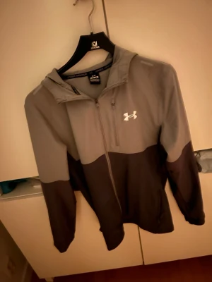 vindjacka Under Armour - Snygg tvåfärgad vindjacka från Under Armour i grått och svart. Jackan har huva, dragkedja framtill och Under Armour-logga på bröstet. Perfekt för blåsiga dagar och sportiga outfits. Tillverkad i lätt och funktionellt material.