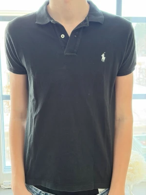 Svart pikétröja från Ralph Lauren - Klassisk svart pikétröja från Ralph Lauren med vit broderad logga på bröstet. Tröjan har krage, korta ärmar och knappar vid halsen. Perfekt till jeans eller shorts för en clean och stilren look.