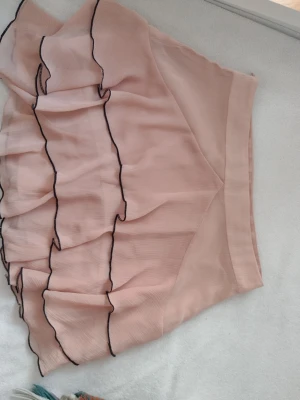 Ljusrosa volangkjol med svarta kanter - Supersöt ljusrosa kortkjol med flera lager volanger och svarta vågiga kanter. Kjolen har en mjuk och luftig känsla, perfekt för dig som gillar en feminin och lekfull stil. Materialet känns som tunn polyester och kjolen har en normal-mini passform. 