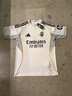 Real Madrid matchtröja Adidas vit - Snygg vit Real Madrid matchtröja från Adidas med svarta detaljer och gula linjer. Tröjan har klubbmärke på bröstet, Adidas-logga och sponsortryck 'Emirates Fly Better' framtill. Helt ny med lappar. 