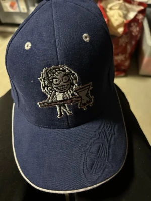 Mörkblå keps med broderad figur - Snygg mörkblå keps med en broderad tecknad figur som håller en elgitarr på framsidan. Kepsen har vita detaljer och en diskret brodyr på skärmen. Perfekt för dig som gillar unik och lekfull stil. Helt ny.