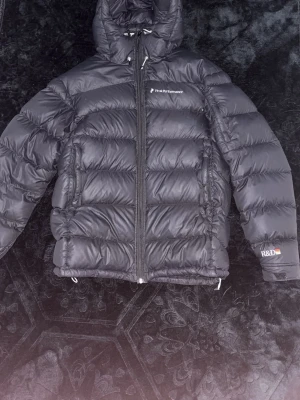 Svart dunjacka från Peak Performance - Säljer en svart dunjacka från Peak Performance med huva och dragkedja framtill. Jackan har en puffig design med breda kanaler och logga på bröstet samt ärmen. Perfekt för kalla vinterdagar och riktigt varm tack vare dunfyllningen.