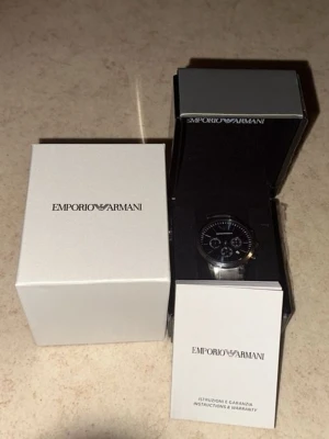 Emporio Armani silverfärgad klocka kan sänka pris VID SNABB AFFÄR. - Stilren klocka från Emporio Armani med svart urtavla och silverfärgat armband i metall. Klockan har kronograf-funktion och tydliga indexmarkeringar. Levereras i originalask med tillhörande manual och garanti.