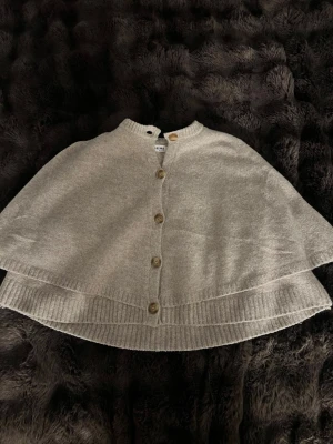 Beige poncho med knappar från missäy - Snygg beige poncho med träknappar framtill och ribbade kanter. Ponchon har en lös och avslappnad passform och är knappt andvänd endast andvänd 1-2 gånger 