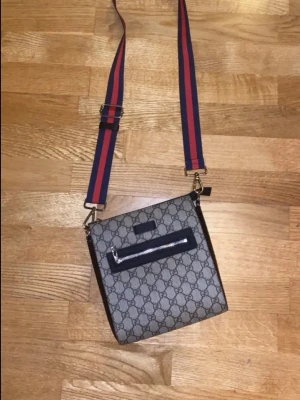 Gucci axelväska - Snygg axelväska från Gucci i beige och mörkbrunt GG-monogram. Väskan har en justerbar axelrem i rött och marinblått med silverdetaljer och en praktisk ytterficka med dragkedja. Perfekt för dig som vill ha en trendig och klassisk accessoar.