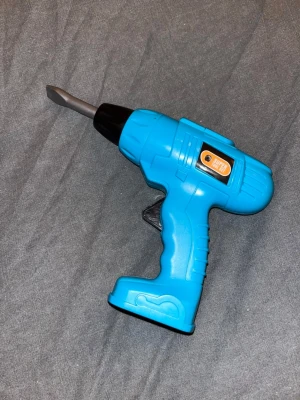 Leksaksborrmaskin i plast - Leksaksborrmaskin i plast med avtagbart grått borrhuvud. Maskinen har en svart knapp vid handtaget- trycker man på den så snurrar borrhuvudet. Perfekt för barn som gillar att leka hantverkare och bygga saker.