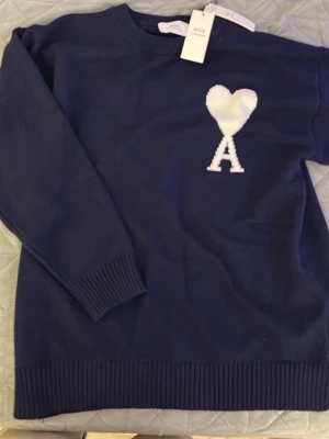 Mörkblå stickad tröja från Ami - Mörkblå stickad tröja från Ami med ett vitt hjärta och bokstaven A broderat på bröstet. Tröjan har rund halsringning, långa ärmar och ribbade muddar vid ärmslut och nederkant. Perfekt för dig som gillar stilrena detaljer.