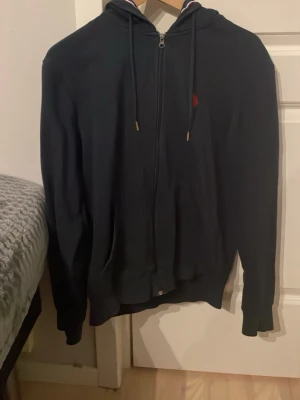 Mörkblå zip hoodie med huva - Snygg mörkblå hoodie med dragkedja och huva. Tröjan har dragsnören i huvan och en liten röd logga på bröstet. Insidan av huvan har en randig detalj i vitt och rött. Perfekt för en avslappnad stil.