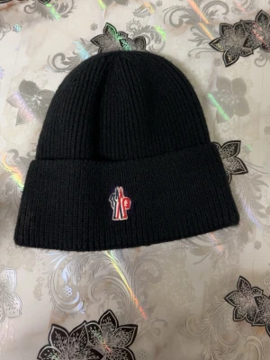 Svart Moncler mössa med broderad logga - Snygg svart ribbstickad mössa från Moncler med uppvikt kant och broderad logga framtill i rött, vitt och blått. Klassisk modell som passar perfekt till kalla dagar. Märkt med modellen ZIND på insidan.