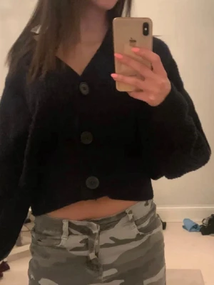 Svart cropped kofta med stora knappar - Säljer en svart, mjuk och cropped kofta med tre stora knappar framtill. Koftan har en djup v-ringning och långa ärmar, perfekt för att skapa en avslappnad och trendig look. Materialet ser ut att vara fluffigt och bekvämt.