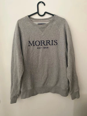 Grå sweatshirt från Morris gents  - Säljer en klassisk grå sweatshirt från Morris med mörkblå broderad logga och texten 'EST 1968' på bröstet. Tröjan har rund halsringning, ribbade muddar och är tillverkad i mjuk bomullsblandning. Perfekt för en avslappnad och stilren look.