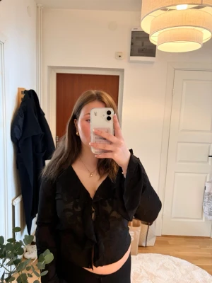 Svart transparent blus med volang - Snygg svart blus med transparent tyg och broderade mönster. Blusen har volangdetaljer längs urringningen och knytning framtill. Ny med prislapp kvar