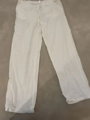 Vita linnebyxor med dragsko - Säljer ett par vita linnebyxor med avslappnad passform och dragsko i midjan. Byxorna har raka ben och två fickor framtill. Perfekta för en chill och stilren look. Materialet är lätt och luftigt linne, vilket gör dem sköna att bära.