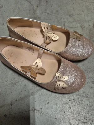 Ballerinaskor - Säljer ett par glittriga ballerinaskor från H&M i storlek 33. Skorna är beige med massor av glitter och har dekorativa guldfärgade fjärilar på ovansidan samt ett elastiskt band över foten. Perfekta för dig som vill ha något extra sött till outfiten.