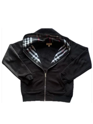 Svart hoodie från Burberry med dragkedja - Svart hoodie från Burberry med dragkedja framtill och klassiskt rutigt foder i huvan. Hoodien har två fickor fram och ribbade muddar vid ärmar och nederkant. Perfekt för en stilren och avslappnad look. Materialet känns mjukt och bekvämt. Passar S men passar XS