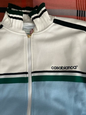 Casablanca blåvit tröja  - Säljer en snygg tröja från Casablanca i ljusblått och vitt med gröna och svarta ränder. Jackan har hög krage, dragkedja framtill och två fickor. Perfekt för dig som gillar sportig och retro stil. HELT NY OCH OANVÖND