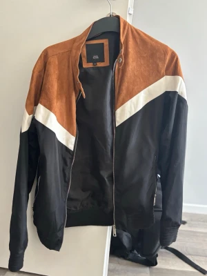Svart och brun jacka med vit detalj - Snygg bomberjacka i svart och brunt med vit diagonal detalj över bröstet. Tight modell. Knappt använd. 