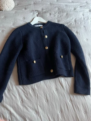 Mörkblå kofta med guldknappar från Zara - Stilren mörkblå kofta från Zara med rund halsringning och fyra dekorativa guldknappar framtill. Koftan har två fickor framtill och är gjord i ett mjukt, stickat material. Perfekt för en trendig och klassisk look.