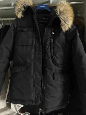Svart parkas från Hollies  - Snygg svart parkas från Hollies med stor huva. Jackan har dragkedja och knappar framtill, flera fickor och justerbar midja. Nypris är 4000kr! Sparsamt använd! Inga hål inga fläckar💕