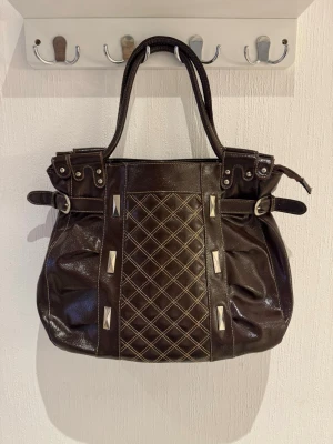 Mörkbrun handväska med nitar - Snygg mörkbrun handväska i glansigt läder med dubbla handtag. Väskan har dekorativa nitar och rutmönster framtill samt spänndetaljer på sidorna. Baksidan har en dragkedjeficka med liten berlock. Perfekt för dig som gillar edgy detaljer.